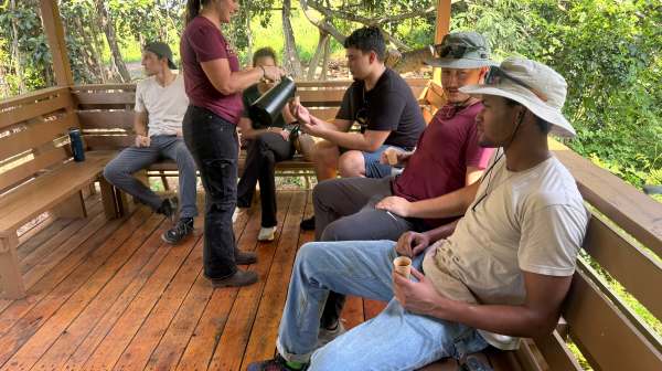 Group of people sitting on a wooden deck, one pouring coffee, surrounded by trees.